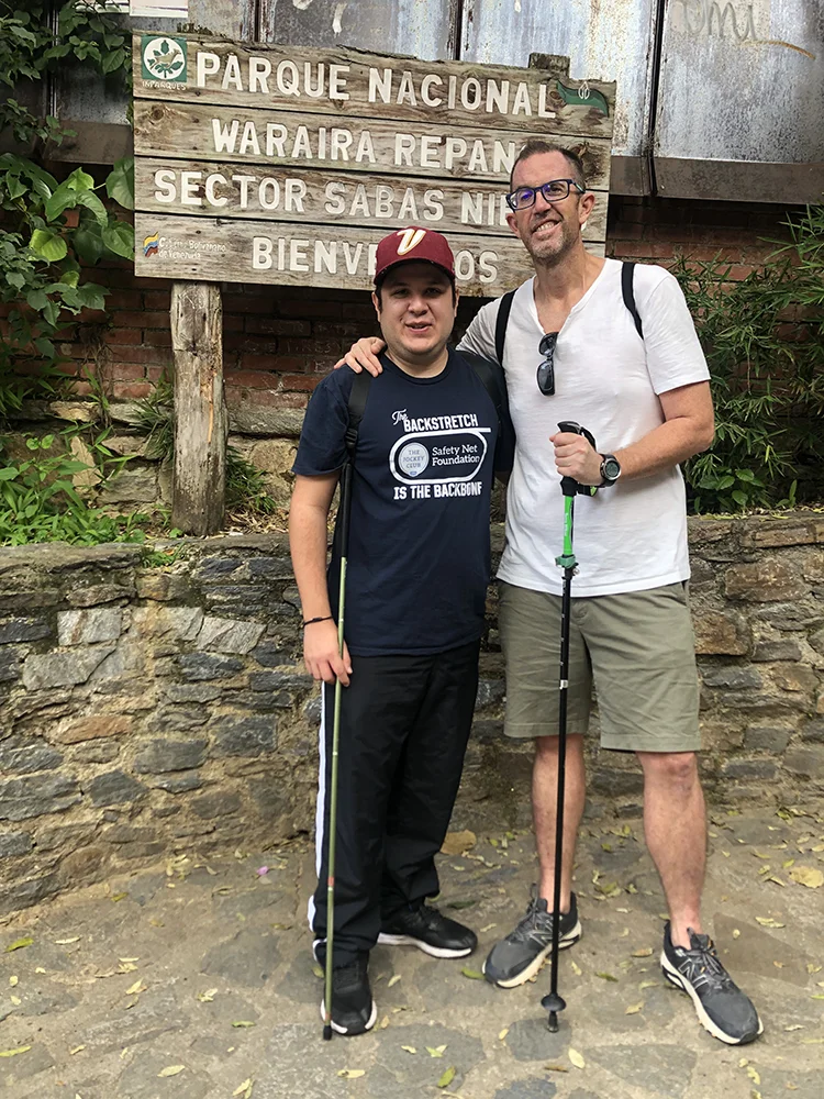 Dos senderistas con bastones frente al cartel de entrada al sector Sabas Nieves del Parque Nacional Waraira Repano, antes de subir al cerro El Ávila