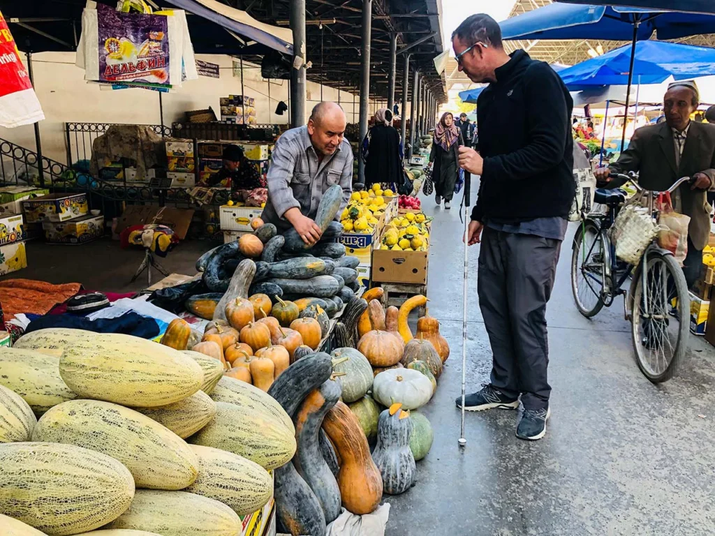 Felipe de pie con su baston blaco y rojo, en un mercado de Margiland observandi unb puesto de frutas junto a los vendedores